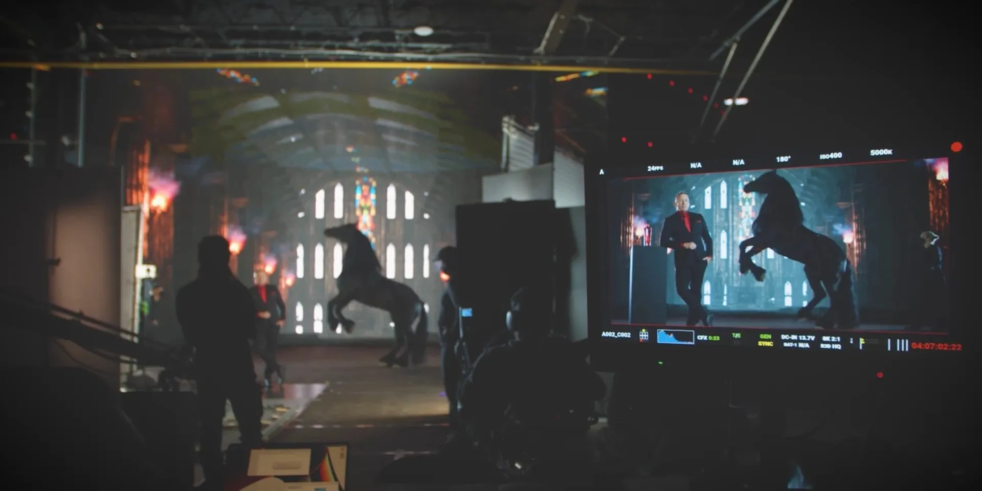 On a warehouse set, crew watch a man in a black suit and a rearing black horse before a cathedral backdrop, the shot also showing on a camera monitor in the foreground.